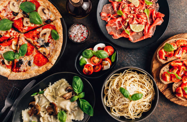 Assortiment de plats italiens comprenant une pizza margherita, des pâtes, des bruschettas, des raviolis aux champignons, des tomates mozzarella et de la charcuterie italienne, tous garnis de basilic frais.